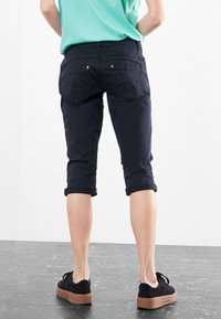 Czarne krótkie spodnie z bawełny, o dopasowanym kroju, z podwiniętymi mankietami i tylnymi kieszeniami. Noszone z zieloną koszulą i czarnymi butami.