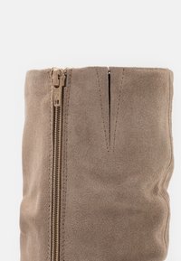 Botte en suédine beige jusqu'au genou avec une fermeture éclair sur le côté, présentant une texture lisse et épurée ainsi qu'un détail de couture verticale sur la partie supérieure.