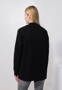 Schwarze gestrickte Strickjacke mit strukturierter Oberfläche, langen Ärmeln und geripptem Saum. Rückansicht, die den lockeren Schnitt und den runden Ausschnitt zeigt.
