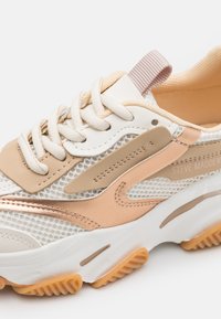 Sneaker mit weißem Mesh-Obermaterial, beigefarbenen Lederakzenten, beigen Schnürsenkeln und einer Gummisohle mit hellorangefarbenen Profil. Markenname angezeigt.