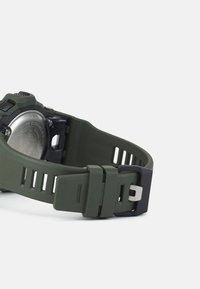 Grønn silikonurrem med teksturert overflate, svart hardware-spenne og utskårne akenter. Metallisk bakdeksel synlig.