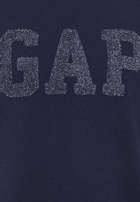 Námořnicky modrá mikina s velkými, texturovanými, třpytivými šedými písmeny tvořícími nápis "GAP". Měkká látka s hladkým, bezvzorem povrchem.