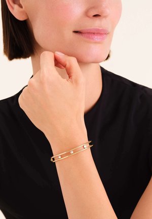 Goldfarbener Metallarmreif mit zwei offenen Bändern, der drei kleine weiße Perlen gleichmäßig verteilt aufweist. Die Hand des Models ruht nahe dem Gesicht und trägt ein schwarzes Oberteil.