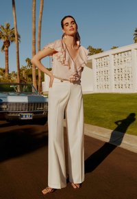Light pink blouse with ruffled neckline, white wide-leg trousers, and strappy brown sandals. Background includes palm trees and vintage car.