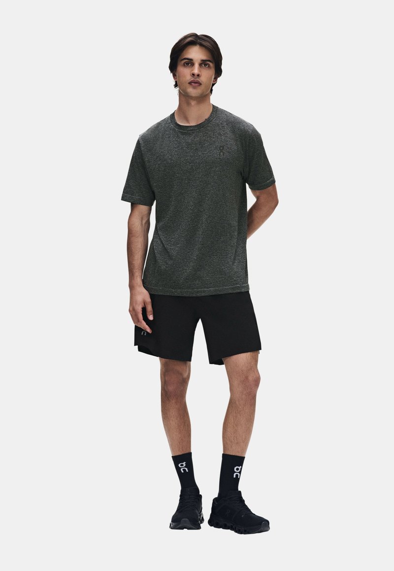 Jeune homme debout portant une chemise à manches courtes gris foncé, un short noir, des chaussettes noires et des chaussures de sport noires sur un fond uni.