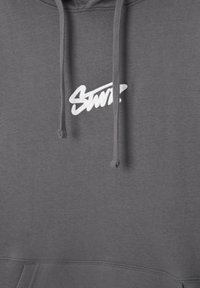 Sweat à capuche gris avec un texte blanc stylisé "Saint" sur la poitrine et des cordons gris assortis pendants à l'avant.