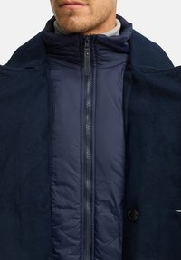 Marineblå dual-layer jakke med et quilted inderlag, lynlås foran og et blødt ydre lag med knapdetalje.