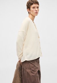 Κρέμ, πουλόβερ με κουμπάκια και χαλαρή εφαρμογή, μακριά μανίκια, φτιαγμένο από μαλακό ύφασμα. Συνδυάζεται με χαλαρές, καφέ παντελόνες.