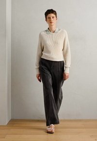 Krema rebrasta pletena majica, svijetlo zelena košulja s kragnom, tamne hlače širokih nogavica i bijele sandale. Model stoji na neutralnoj pozadini.