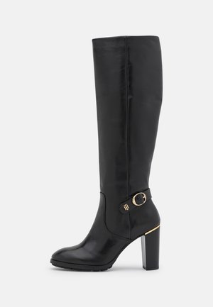 Botte en cuir noire montant jusqu'au genou avec talon bloc, sangle latérale décorative, boucle dorée, et un discret accent doré sur le talon.