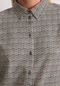 Chemise à boutons présentant un motif géométrique noir et crème, avec un col et cinq boutons sur le devant. Texture lisse.