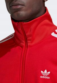 Červená zipová bunda Adidas s vysokým límcem, bílými třemi pruhy a malým bílým logem na hrudi. Hladká, texturovaná tkanina.