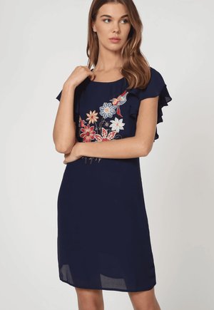 Mujer con un vestido azul marino con mangas con volantes y bordados coloridos de flores y pájaros en el pecho, de pie con los brazos cruzados.