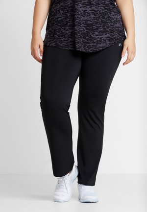 Personne portant des leggings noirs, un haut foncé à motifs et des chaussures de sport blanches, debout devant un fond clair uni.