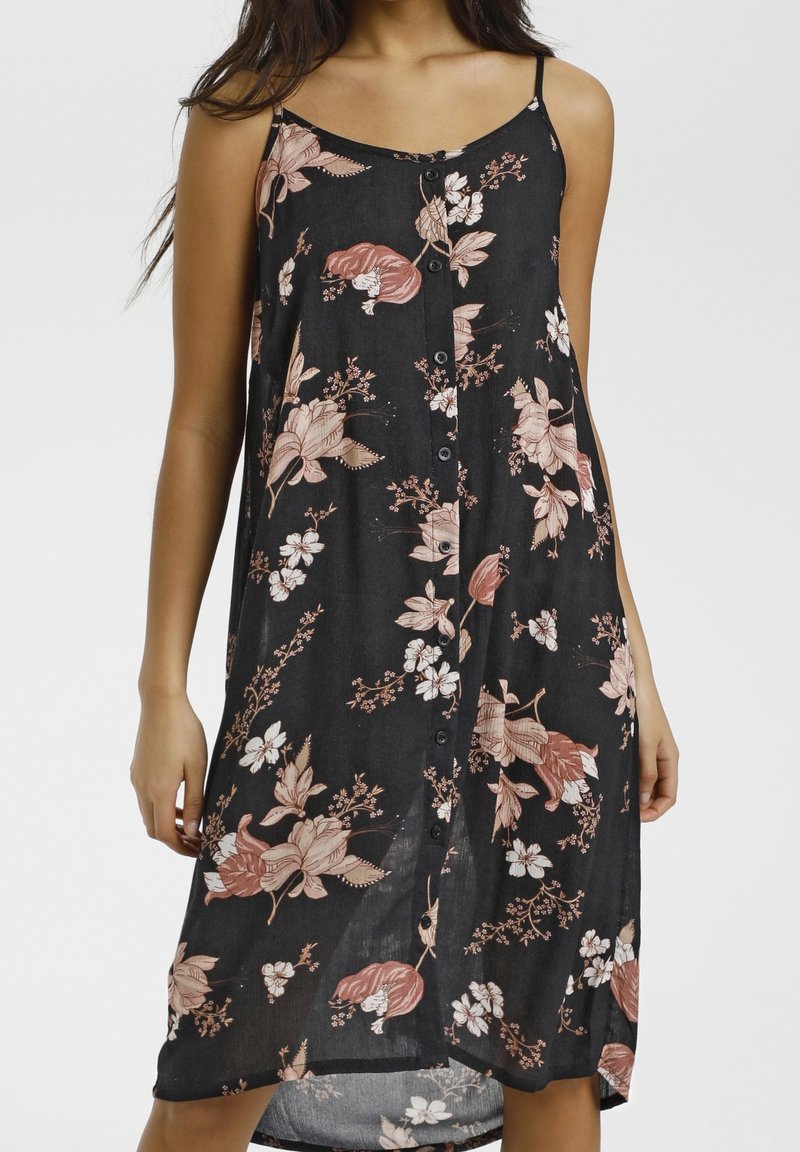 Femme portant une robe noire sans manches avec des imprimés floraux roses et blancs et des boutons à l'avant, debout devant un fond uni.