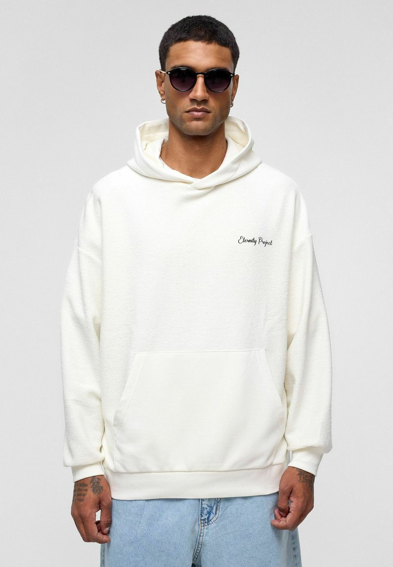 Crème hoodie met textuurstof, voorzak en geborduurd "Eternity Project" logo. Gedorven met denim shorts en een zonnebril.