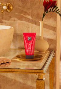 Rød tube med pisket kroppskrem med svart bunn, plassert på et gullfat. Har en teksturert beige bakgrunn og en myk brun håndkle i nærheten.