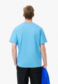 Světle modré bavlněné tričko s krátkými rukávy a kulatým výstřihem, z pohledu zezadu. V rukou má černé kalhoty a modrý ručník.