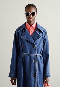 Denim trenchcoat med stor krage, dobbeltknappede knapper, belte i midjen og lange ermer. Båret over en rosa skjorte, accessorert med hvite solbriller.