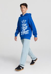 Niebieska bluza z kapturem z białym napisem w stylu graffiti, jasnoniebieskie dżinsy i czarne trampki z białymi akcentami, idące na neutralnym tle.