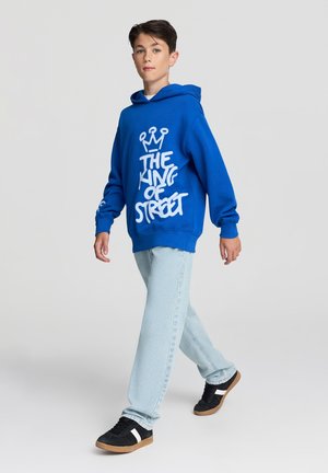 Felpa con cappuccio blu con testo in stile graffiti bianco, jeans azzurri chiari e sneakers nere con dettagli bianchi, mentre cammina su uno sfondo neutro.