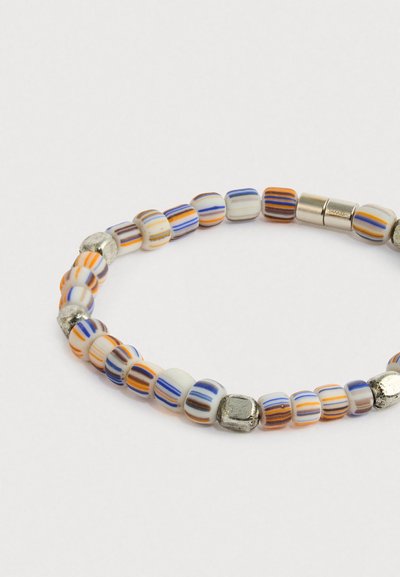 Rugalmas karkötő kis fehér gyöngyökkel, narancssárga, kék és barna csíkokkal, közöttük fémes ezüst színű, kocka alakú gyöngyökkel.