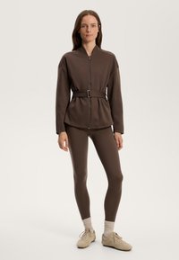 Brun zip-up jacka med midjebälte, i kombination med matchande leggings. Mjuk textur, avslappnad passform och långa ärmar. Jordnära design.