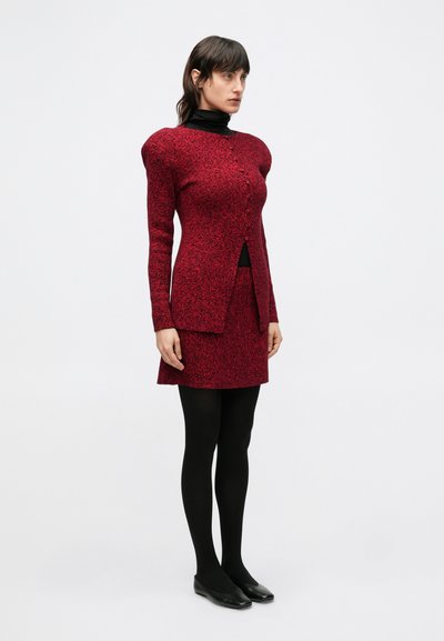 Červené pletené šaty s dlhými rukávmi, so zapínaním na gombíky vpredu, s rázporkom a priliehavým strihom, kombinované s čiernymi pančuchami a balerínkami.