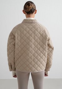 Beige quiltet jakke med diamantmønster, med krage og raglanermer. Glatt tekstur og kontrasterende lys dekor langs sømmen.