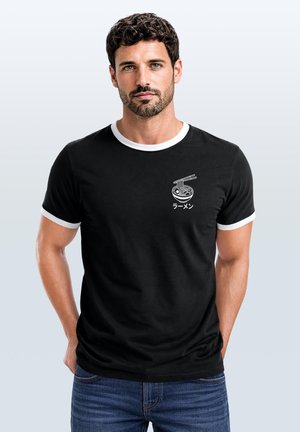 Mann mit dunklen lockigen Haaren und Bart, der ein schwarzes T-Shirt mit weißer Einfassung und einer kleinen Grafik einer Ramen-Schüssel und Essstäbchen auf der Brust trägt.