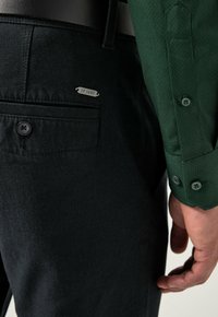 Pantaloni neri texture con tasca posteriore e dettaglio a bottone, abbinati a una camicia verde con motivo e polsini con bottoni. Etichetta "IP 1880" visibile.