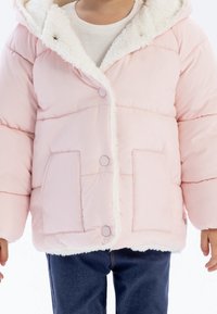 Barn iført en lyserød, polstret jakke med hvid fleece-foering, trykknapper, frontlommer og mørkeblå jeans foran en hvid baggrund.