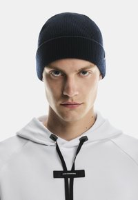 Young man wearing a dark blue knit beanie and a white hoodie with black drawstrings labeled "Swiss Engineering," facing forward.