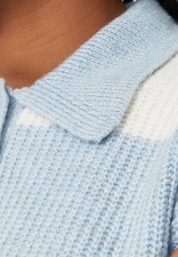 Primo piano di un maglione a collo in maglia azzurro chiaro con un motivo testurizzato e una toppa bianca vicino alla spalla.