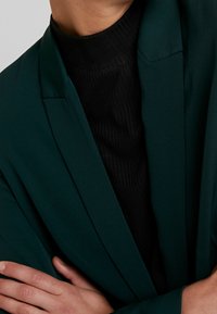 Zelený blazer se strukturovanými ramenními vycpávkami, nošený přes černý žebrovaný rolák. Hladká látka s minimalistickým designem, ruce zkřížené před sebou.