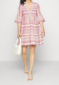 Robe à rayures blanches et roses avec un col en V et des manches évasées. La robe présente des motifs géométriques et une coupe ample et fluide. Sac blanc porté.