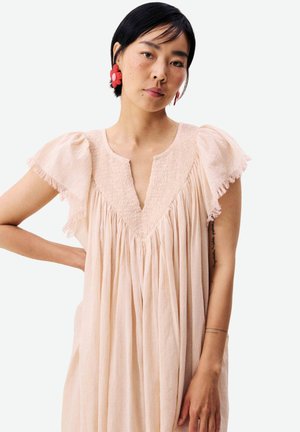 Femme portant une robe fluide rose clair avec des manches à volants et des boucles d'oreilles en forme de fleurs rouges, debout, un bras plié.