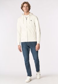 Giovane uomo che indossa una felpa bianca con zip, jeans blu e sneakers bianche mentre è in piedi contro uno sfondo chiaro e uniforme.