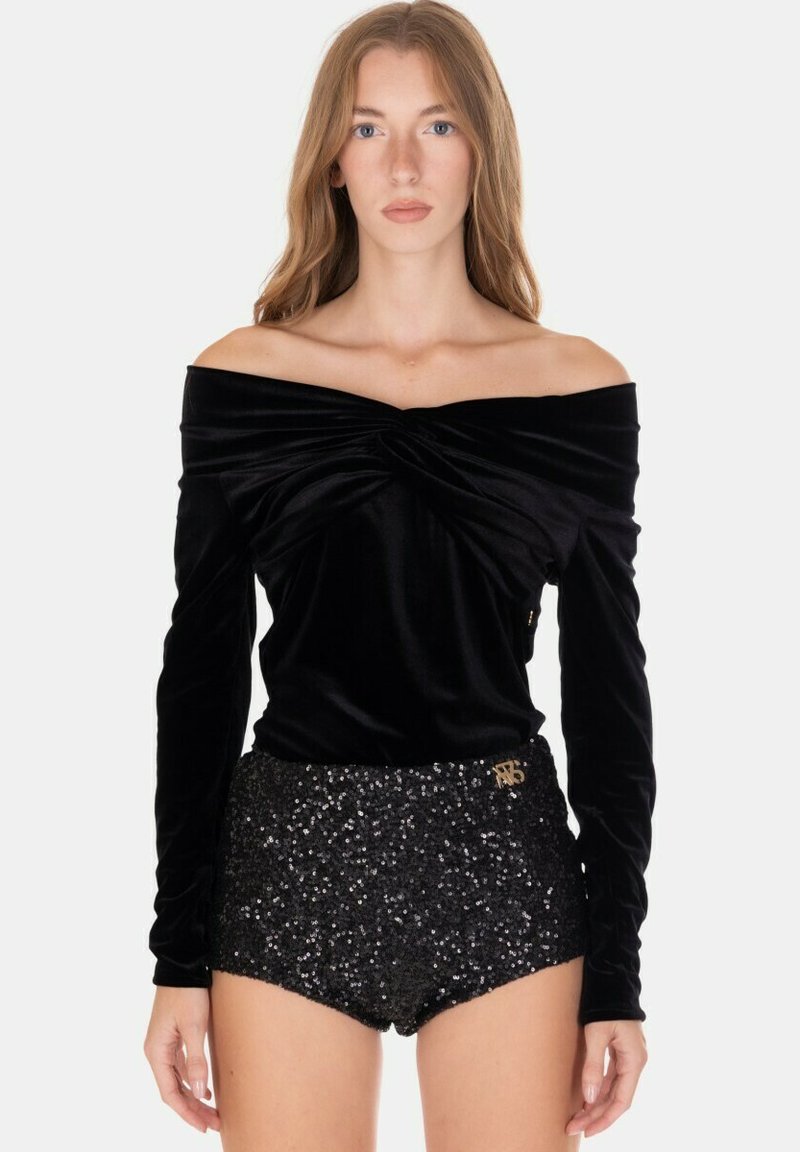 Donna che indossa un top nero in velluto con maniche lunghe e scollatura a barca, abbinato a pantaloncini neri a vita alta con paillettes, su uno sfondo semplice.