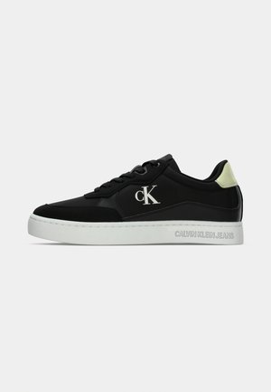 Schwarzer Sneaker mit glattem Obermaterial aus Synthetik, weißer Gummisohle und grünem Akzent an der Ferse. Verfügt über ein Logo an der Seite und den Markennamen auf der Sohle.