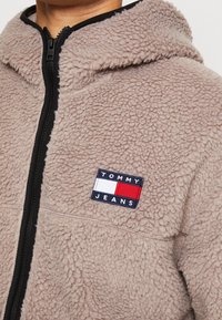 Puszysta beżowa polarowa kurtka z czarnym zamkiem i wykończeniem. Posiada prostokątną naszywkę z logo "TOMMY JEANS" w kolorach granatowym, czerwonym i białym.