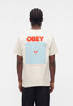 Muž s vrkočmi nosi biele tričko s nápisom „OBEY“ v červených a modrých trojuholníkových líniách okolo malej tváre, text znie „LOVE IN A VOID."