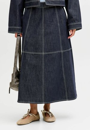 Jupe longue en denim foncé avec des surpiqûres contrastantes, associée à des ballerines beige à lacets, une veste foncée et un sac à main gris tenu dans la main gauche.