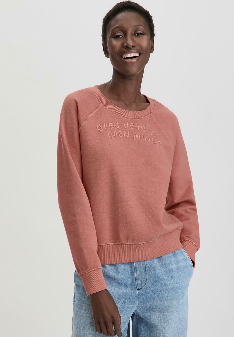 Roze sweatshirt met raglanmouwen, voorzien van verhoogde borduursel tekst "OPEN HEARTS OPEN MINDS." Gecombineerd met lichtblauwe spijkerbroek.