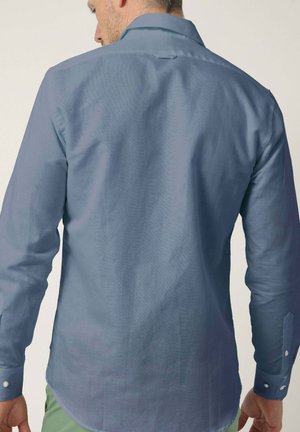 Hombre con una camisa azul de manga larga, abotonada, visto desde atrás, con las manos relajadas a los lados.