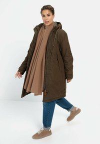 Lange olivgrüne isolierte Jacke mit Kapuze, beiger Strickschal, Jeans und hellbraunen Sneakers. Sie verfügt über Reißverschlüsse und strukturierte Stoffe.