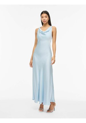 Kobieta stojąca w bezrękawnikowej, jasnoniebieskiej sukience z satyny z dekoltem typu kaskada i sandałach na wysokim obcasie na białym tle.