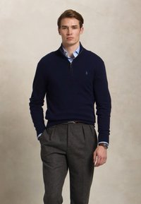 Marineblauwe trui met ritsbijlage, gedragen over een lichtblauw overhemd met kraag. Donkergrijze geplooide broek maakt de outfit compleet. Eenvoudig logo op de borst.