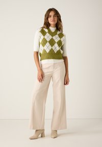 Grünes und weißes Argyle-Weste über einem kurzärmeligen weißen Oberteil, kombiniert mit hellbeigen, weit geschnittenen Hosen und hellbeigen, hohen Stiefeln.