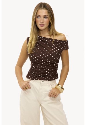 Femme aux longs cheveux blonds portant un haut marron à pois dévoilant les épaules, un pantalon large crème, des bracelets dorés et beiges, ainsi que des bagues.